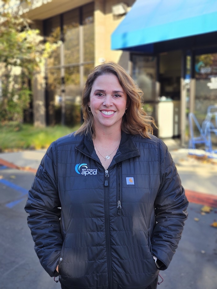 Woman standing outside in blue jacket with SLO County Air Pollution District Logo on it smiling.