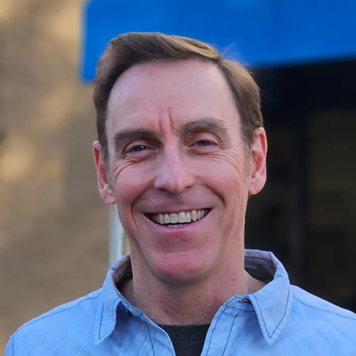 A smiling man with short brown hair in a blue shirt stands outside in front of a blurred building and blue awning.