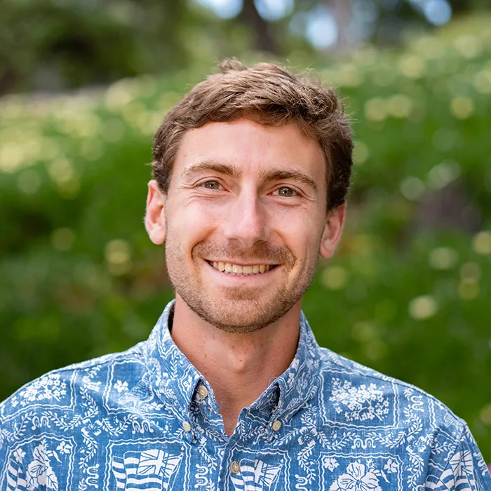 Smiling man in a blue patterned shirt stands outdoors with greenery blurred in the background.