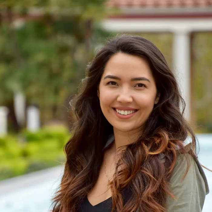 Smiling woman with long wavy hair stands outdoors in front of greenery and a building.