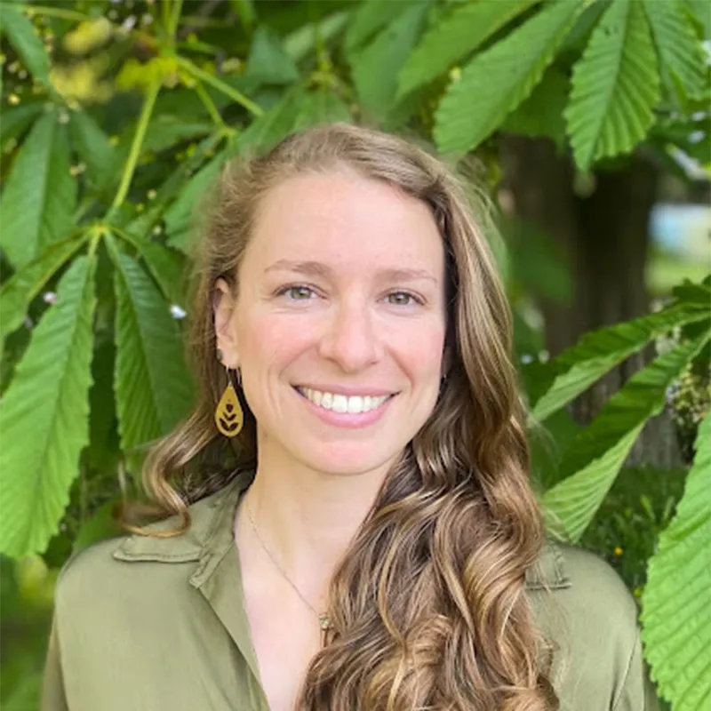 Smiling woman with long wavy hair stands in front of large green leaves, wearing a green shirt and leaf earrings.