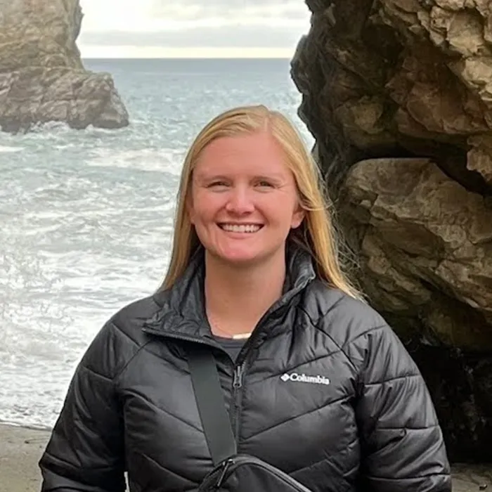 Smiling woman in a black jacket stands by rocky cliffs with the ocean in the background.