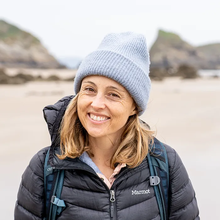 Woman in a gray beanie and puffer jacket smiling on a sandy beach with rocky cliffs in the background.