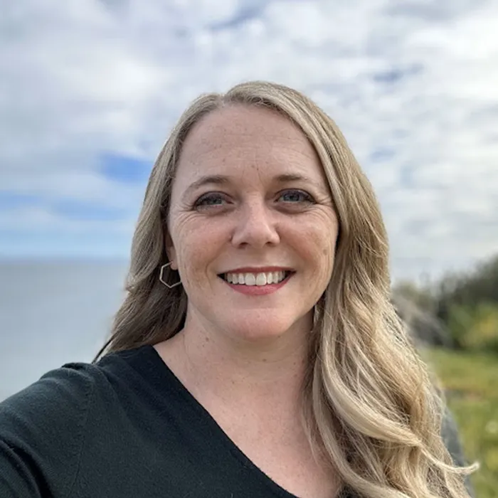 Smiling woman with long blonde hair stands outdoors with a cloudy sky and greenery in the background.