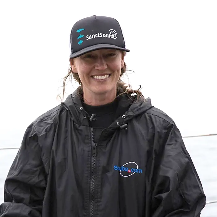 Smiling person in a black jacket and cap, standing outdoors with a white background.