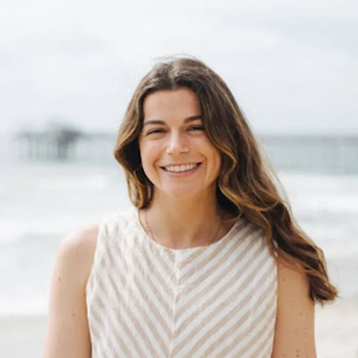 A woman with long brown hair smiles at the beach, wearing a sleeveless striped top.
