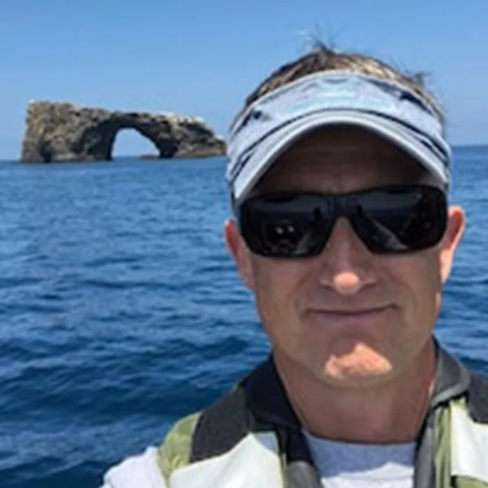 A man wearing sunglasses and a visor takes a selfie on the ocean with a rock arch in the background.
