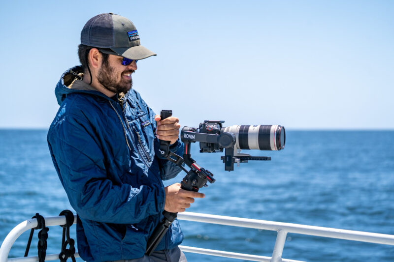 Man on a boat out on the ocean holding professional camera gear and appears to be photographing something in the ocean.