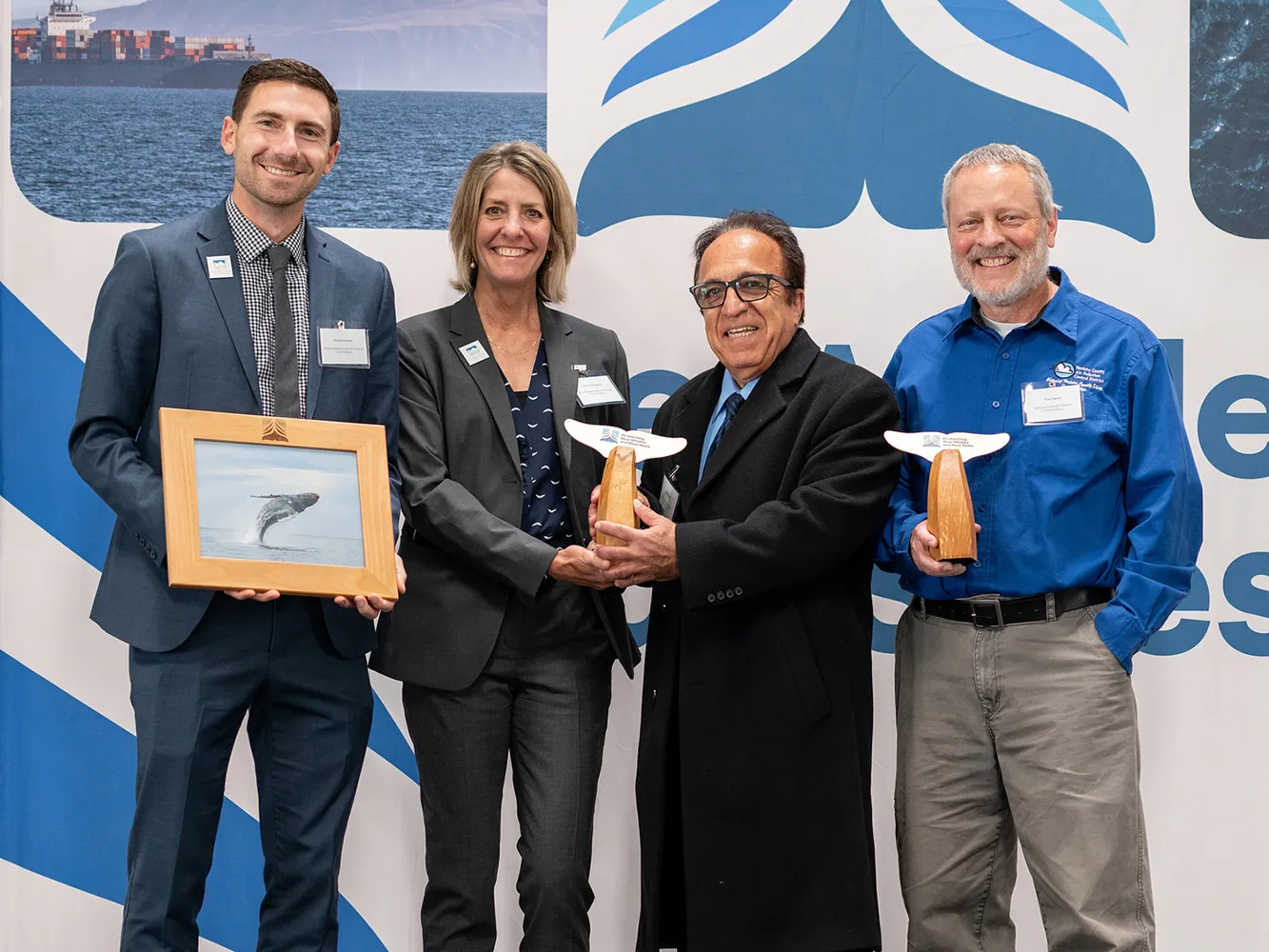 Four people smiling at an event; two hold whale tail trophies, one holds a framed whale photo.