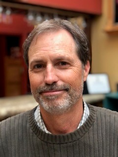 Smiling middle-aged man with short brown hair and beard, wearing a brown sweater indoors.