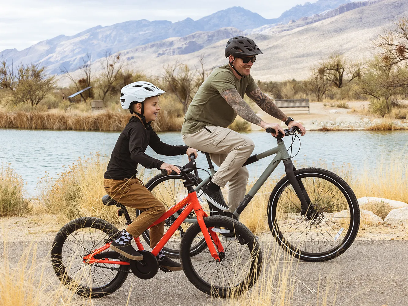 A man and a child ride bikes together on a path by a lake, both wearing helmets and smiling.