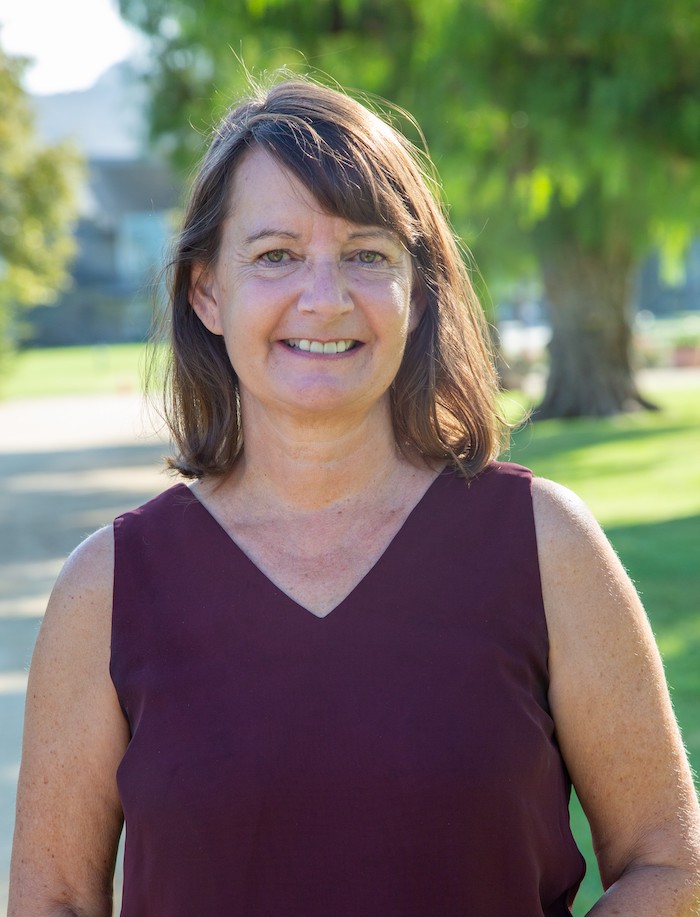 A woman with brown hair smiles outdoors, wearing a sleeveless maroon top, with trees in the background.