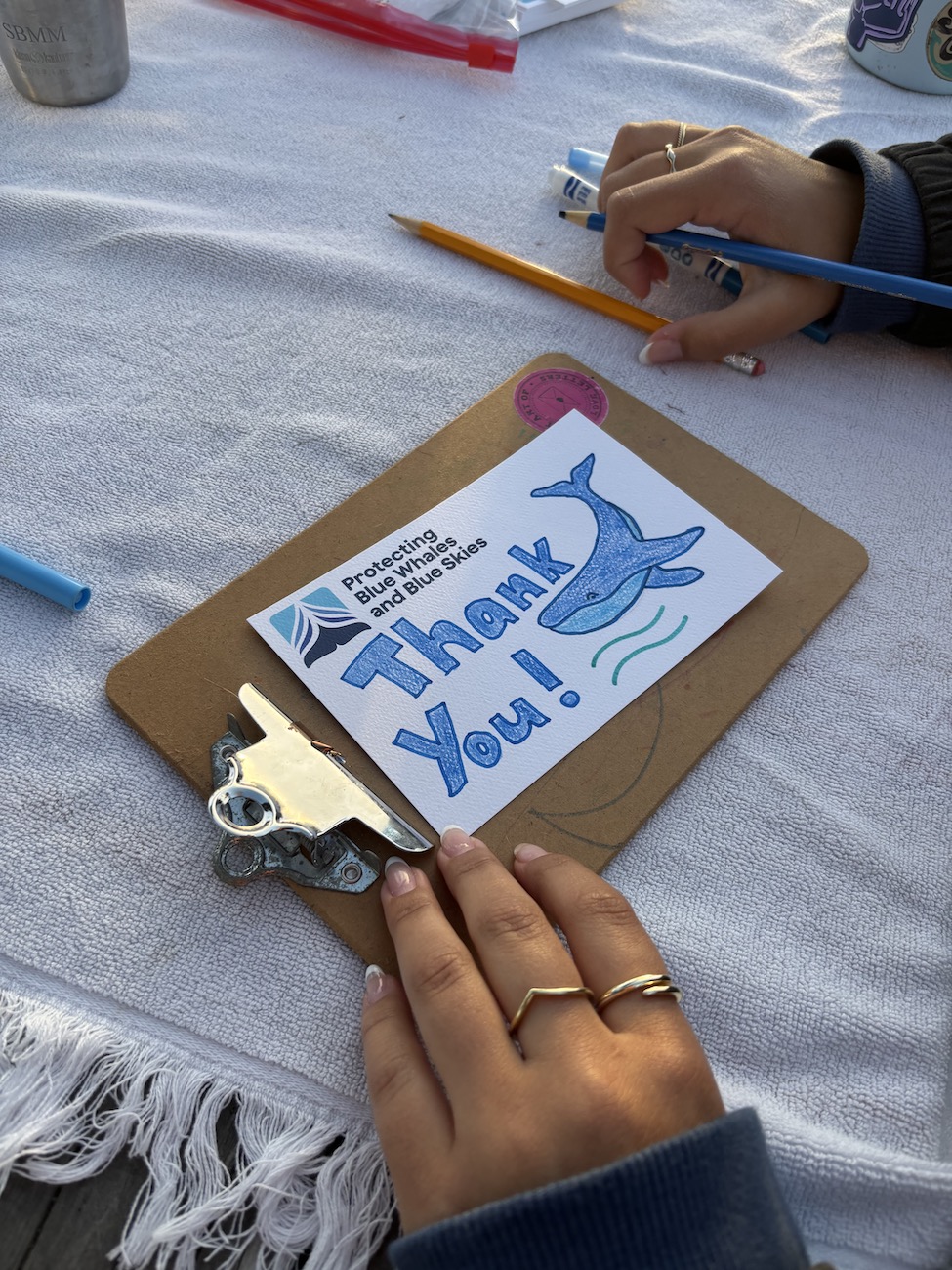 A clipboard holds a postcard that someone is drawing to say "thank you" with a picture of a whale and the blue whales and blue skies logo featured.