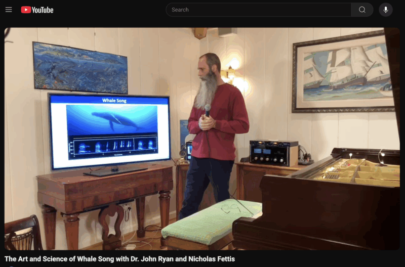 John Ryan stands with microphone looking at a screen depicting a whale and acoustic recordings