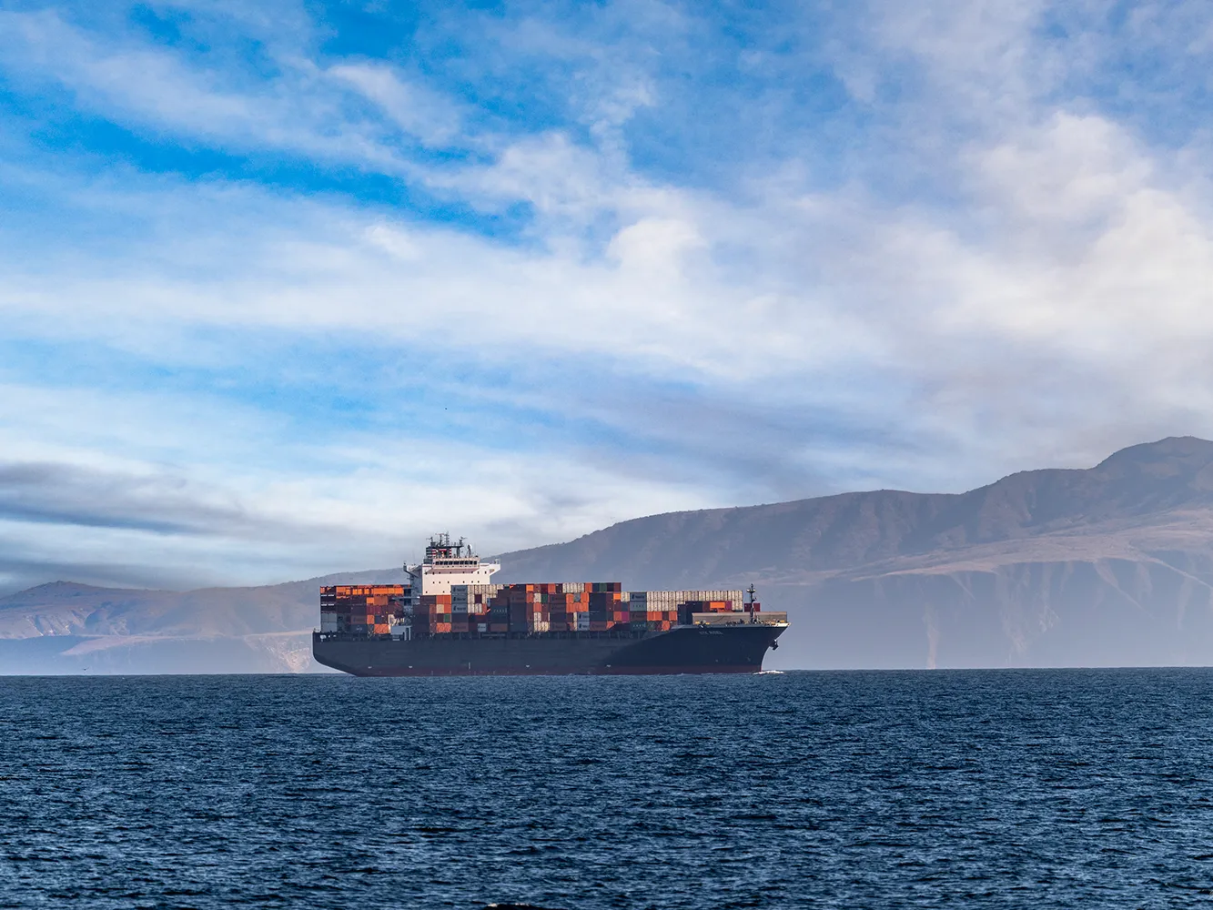 Noise 008_©CLEVENGER-241122 A large cargo ship carrying containers sails on the ocean with mountains and clouds in the background.