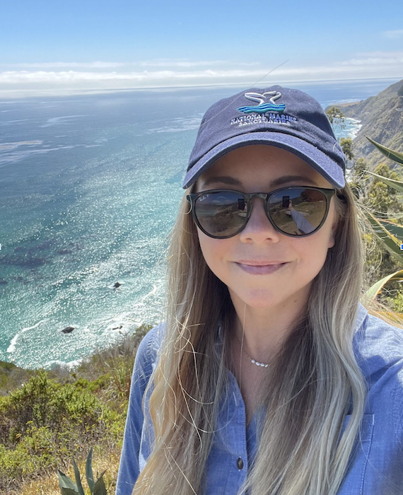 Woman in sunglasses and a hat smiles from atop an island with ocean behind. Laura