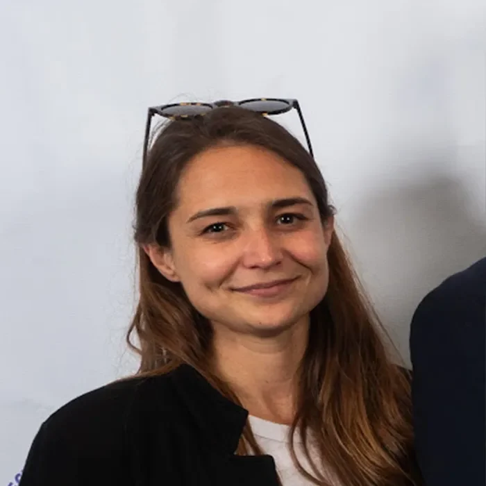 A woman with long brown hair, smiling, wearing glasses on her head, and a black jacket against a white background.