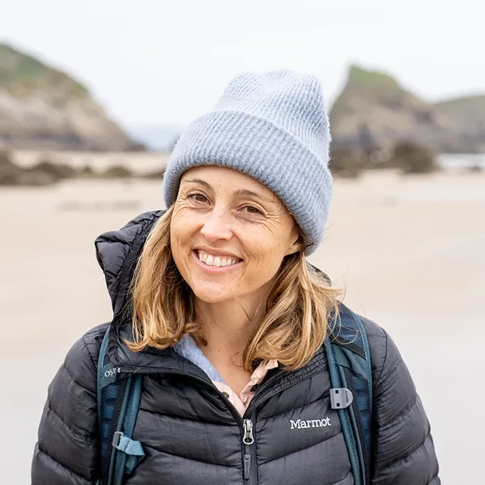 Woman in a gray beanie and puffer jacket smiling on a sandy beach with rocky cliffs in the background.