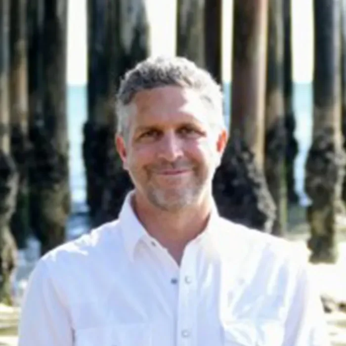 Smiling man in a white shirt stands on a beach with tall wooden pillars in the background.