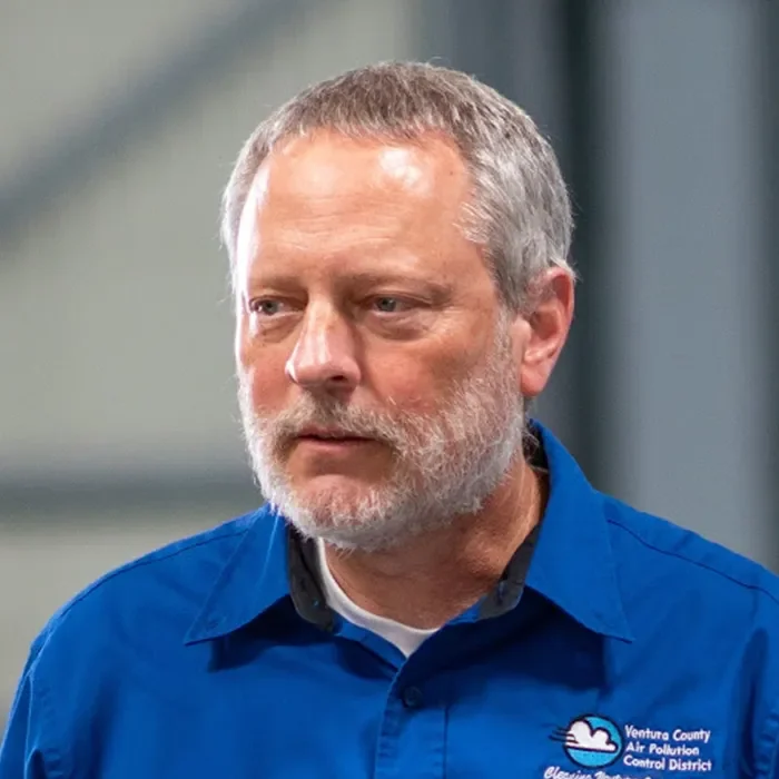 A man with gray hair and beard wears a blue shirt with a Ventura County Air Pollution logo.