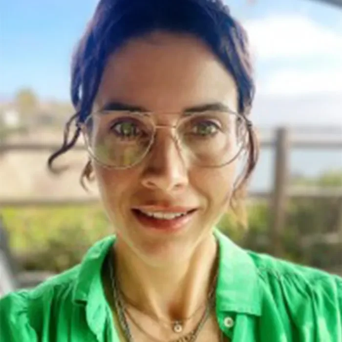 About becca tucker Woman with glasses and dark hair, wearing a green shirt, smiling outdoors with a blurred background.