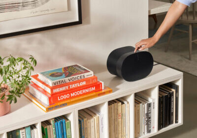 A person presses a button on a black Sonos speaker placed on a shelf with books and a green plant.