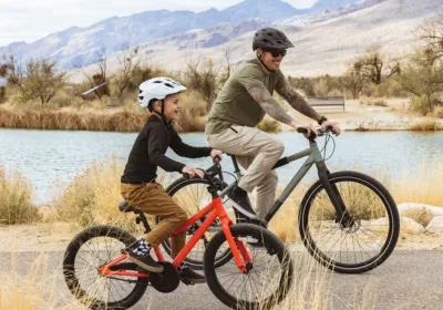 An adult and a child ride bikes together on a path by a lake, wearing helmets, with mountains in the background.
