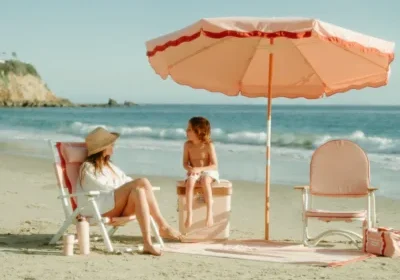 A woman and child sit under a pink umbrella on a sandy beach, with the ocean in the background.
