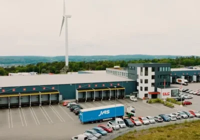 A logistics warehouse with loading docks, parked cars, a blue JAS truck, and a wind turbine in the background.