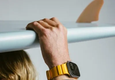 A person with a yellow smartwatch holds a surfboard above their head, fin visible in the background.
