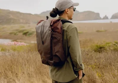 Person wearing a gray hat and sunglasses, standing outdoors with a large brown backpack, facing a scenic landscape.