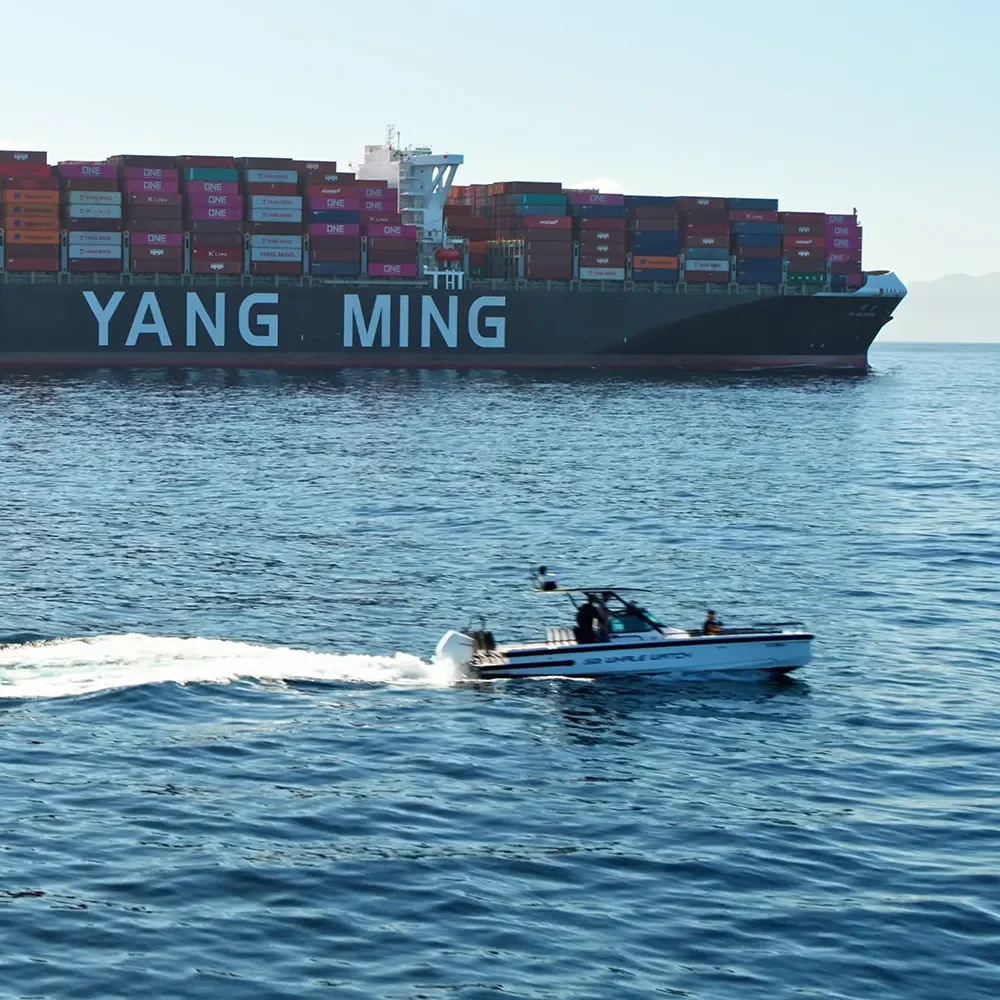 A small motorboat passes in front of a large Yang Ming container ship on the open water.