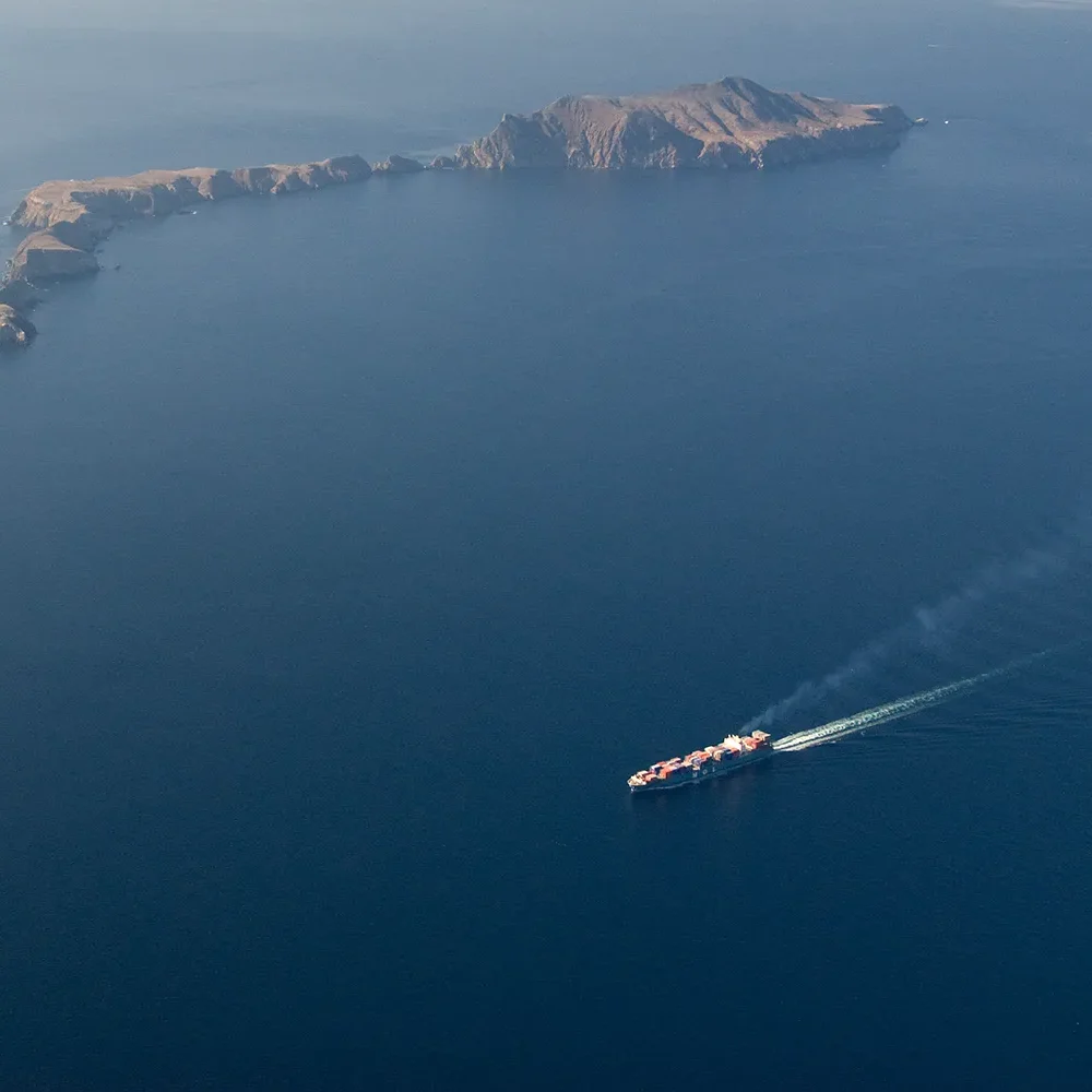 A cargo ship sails near a rocky island in a vast blue sea, leaving a visible wake behind it.