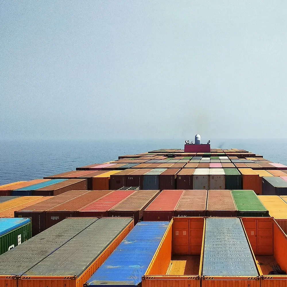 A cargo ship carrying colorful shipping containers sails across a calm, open sea under a hazy sky.