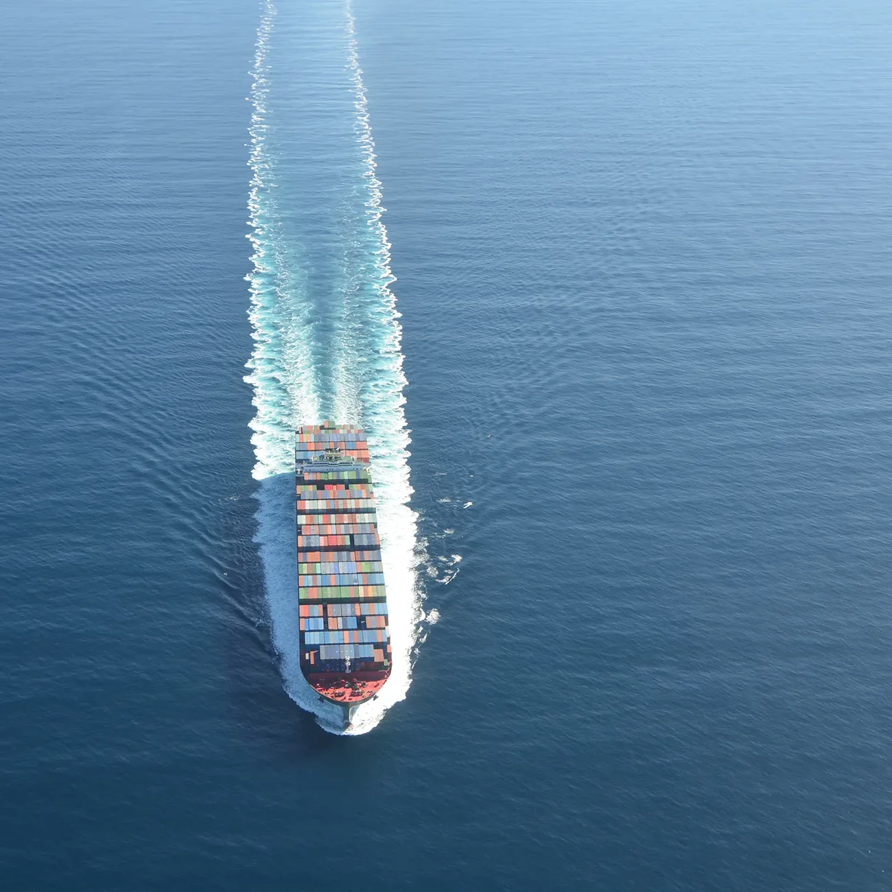 A cargo ship sails through calm blue ocean water, leaving a visible wake behind it.