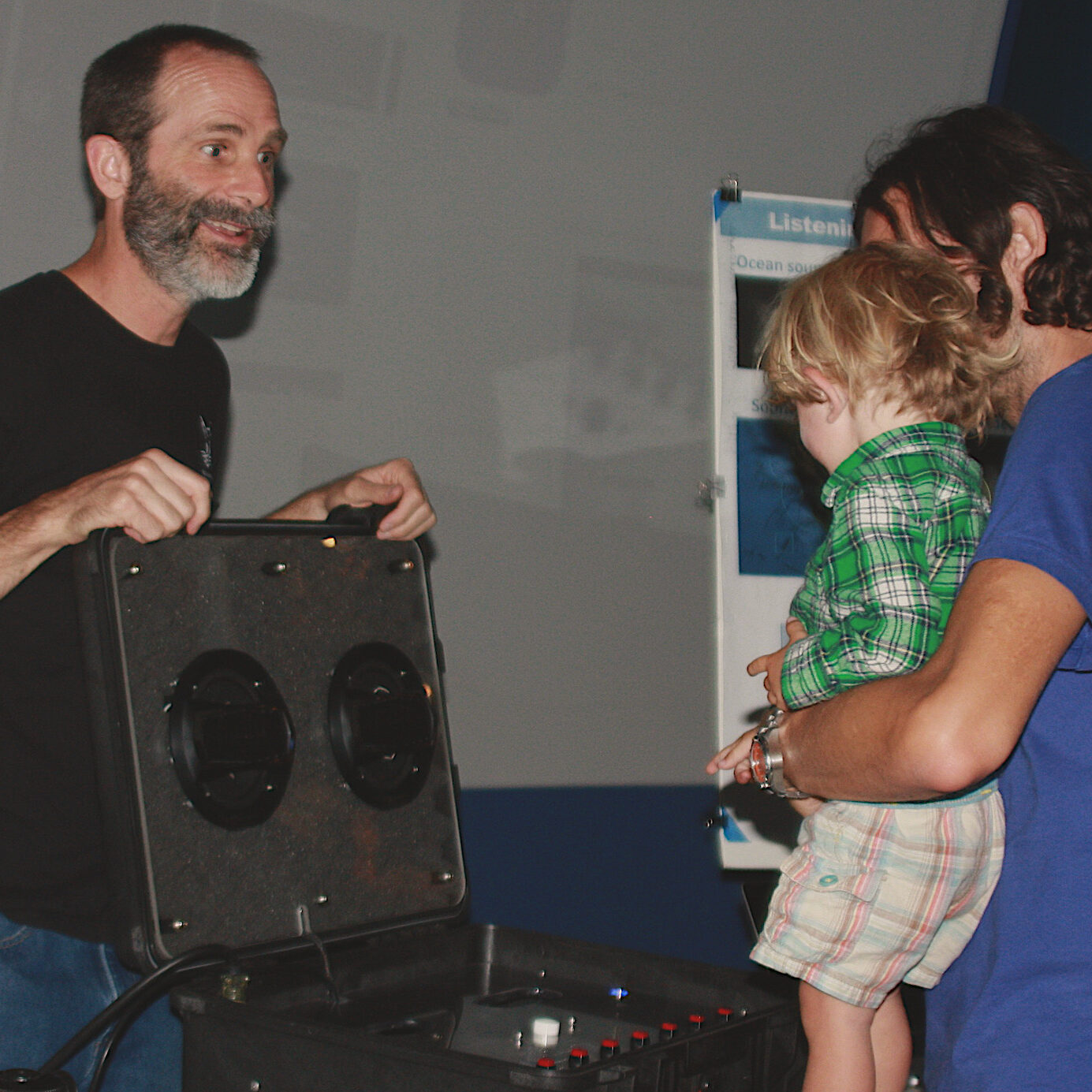 ryjo_exhibit (1) John Ryan stands with a speaker set in front of a child held by an adult who appear to be listening.