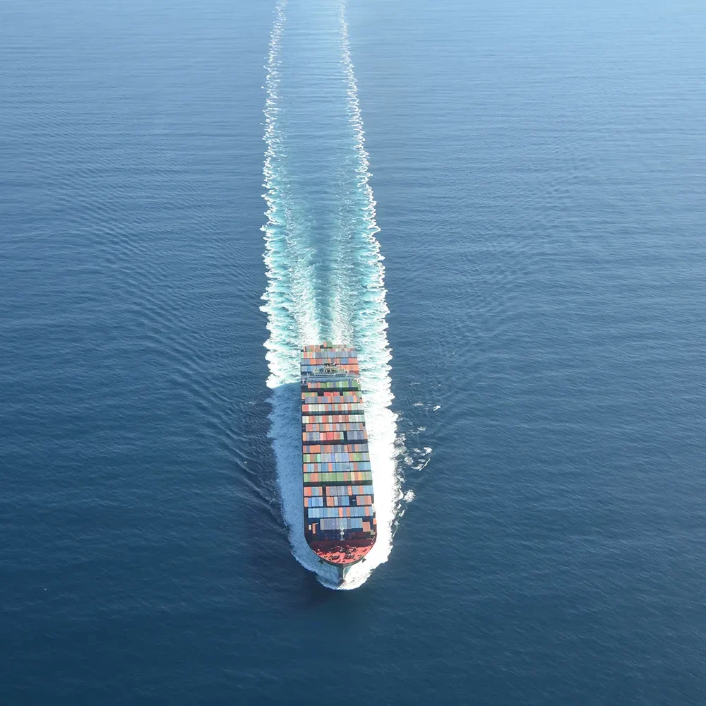 A cargo ship sailing through calm blue ocean water, leaving a white wake behind.