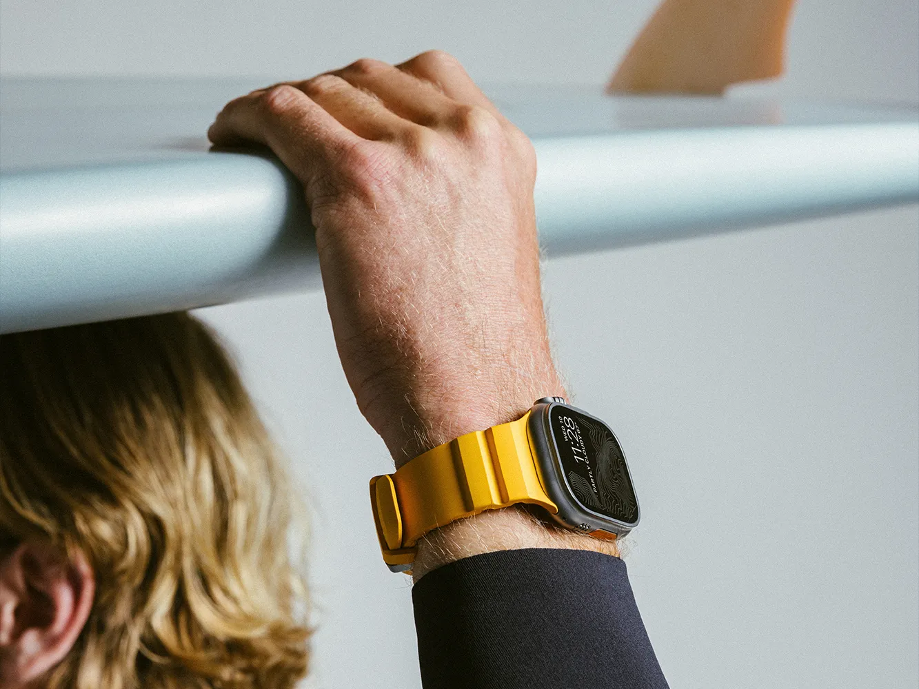 Person with blond hair holding a surfboard, wearing a smartwatch with a yellow band on their wrist.