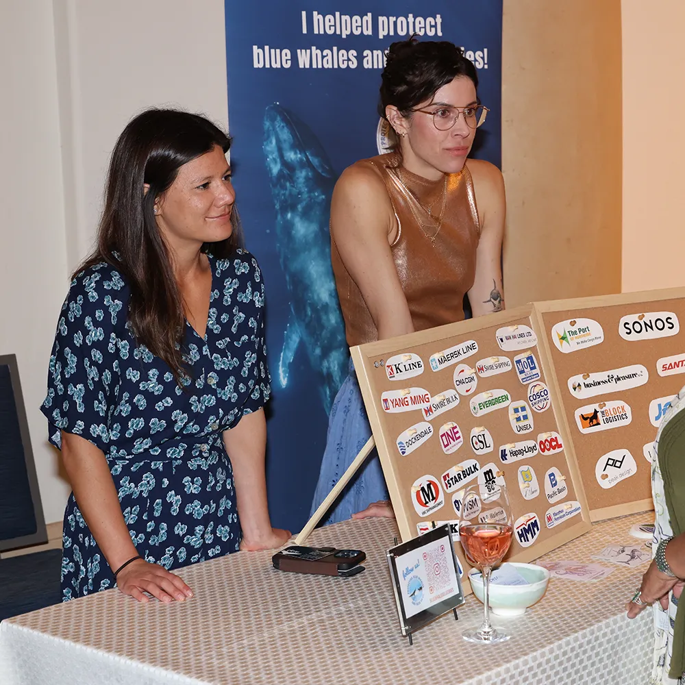 Two women stand behind a table with sponsor logos at an event, a whale conservation poster behind them.