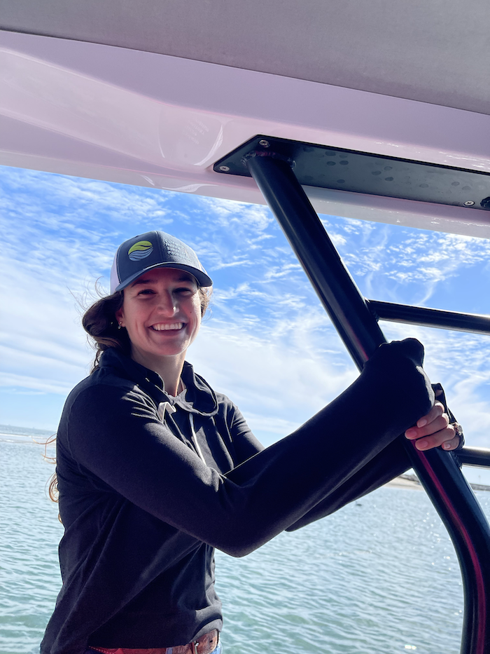 Woman in a baseball hat smiling on a boat with ocean and blue sky behind.