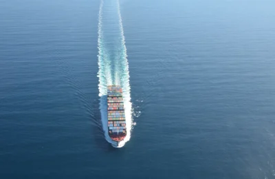 A cargo ship sails through calm blue ocean water, leaving a visible wake behind it.