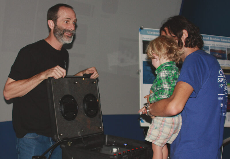 John Ryan stands with a speaker set in front of a child held by an adult who appear to be listening.