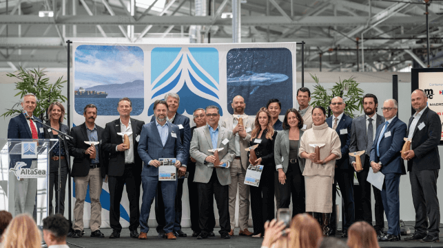 A group of people pose on stage holding awards at an indoor event with a blue and white backdrop.