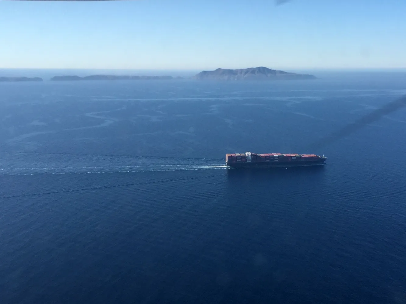 vsr-Copy of Ship Islands - Jess A large cargo ship sails across calm blue ocean water, with an island visible in the distance.
